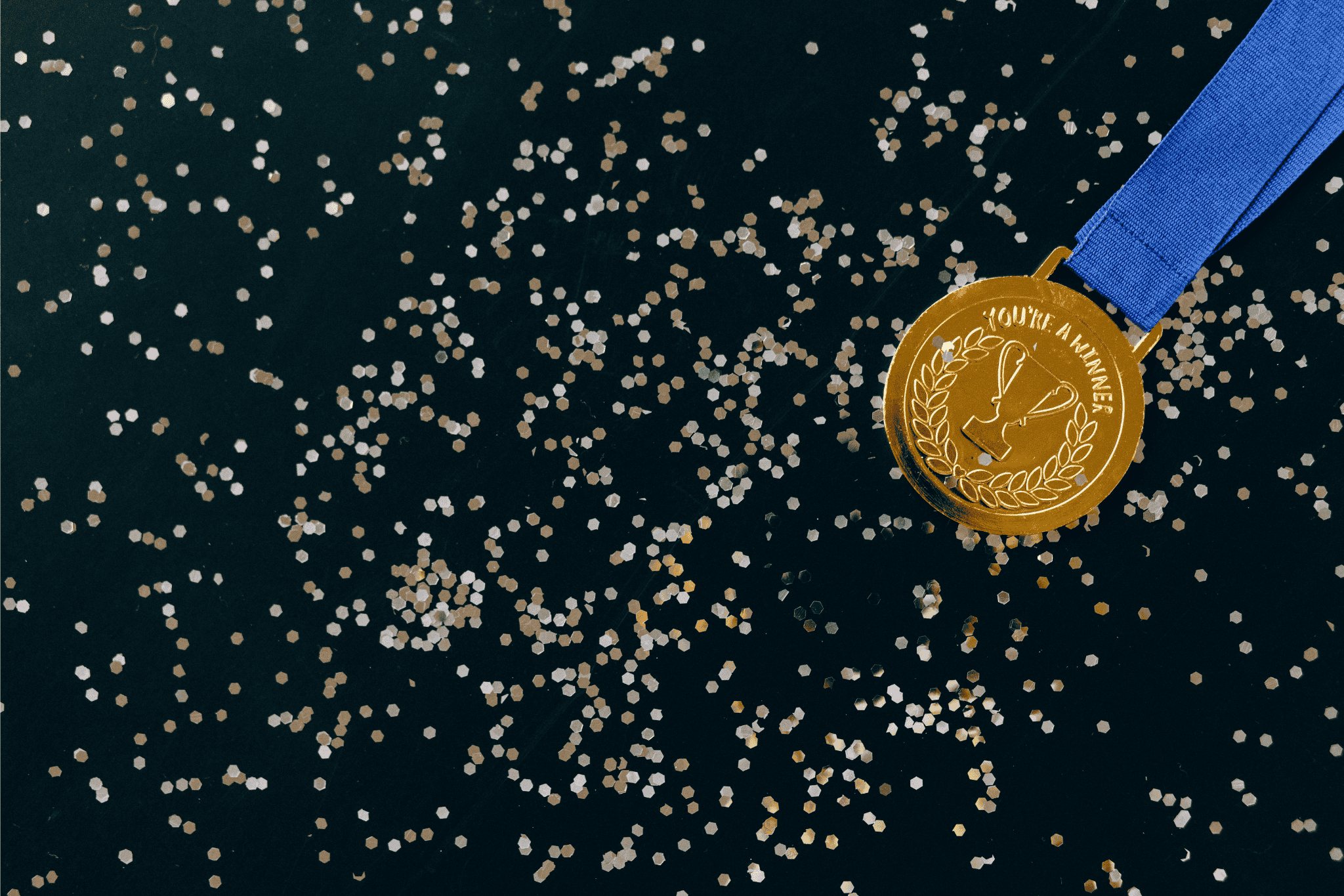 Gold medal with blue ribbon on a dark surface covered in confetti with the words 'You're a Winner' engraved