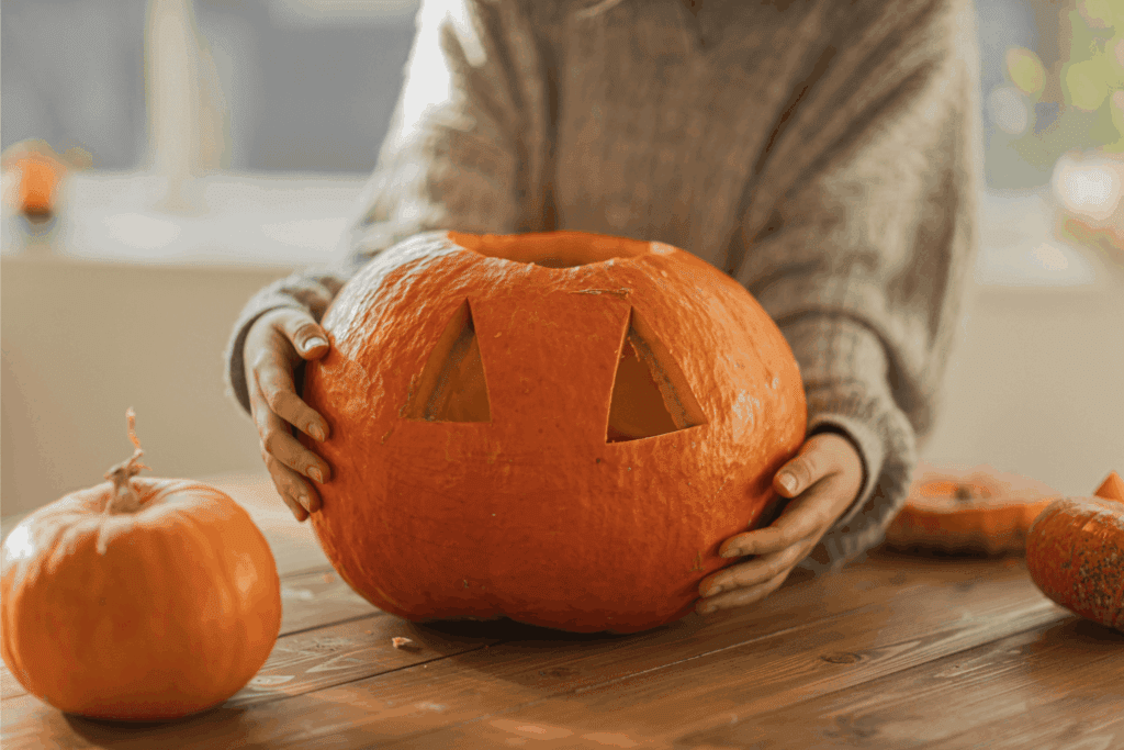Person carving a Halloween pumpkin jack-o’-lantern with triangular eyes on a wooden table.