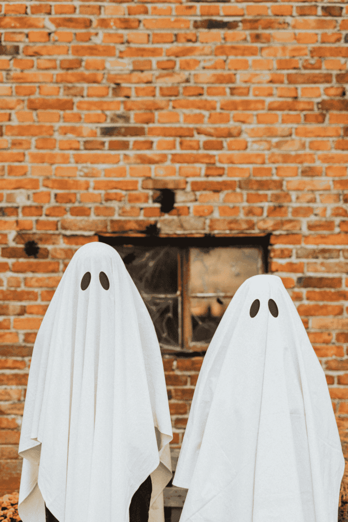 Two people in ghost costumes with white sheets and black eyes standing in front of a brick wall for Halloween.