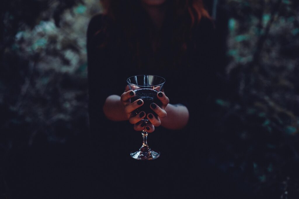 A person dressed in dark clothing holds a gothic-style glass goblet filled with a dark liquid, evoking a mysterious and eerie atmosphere.