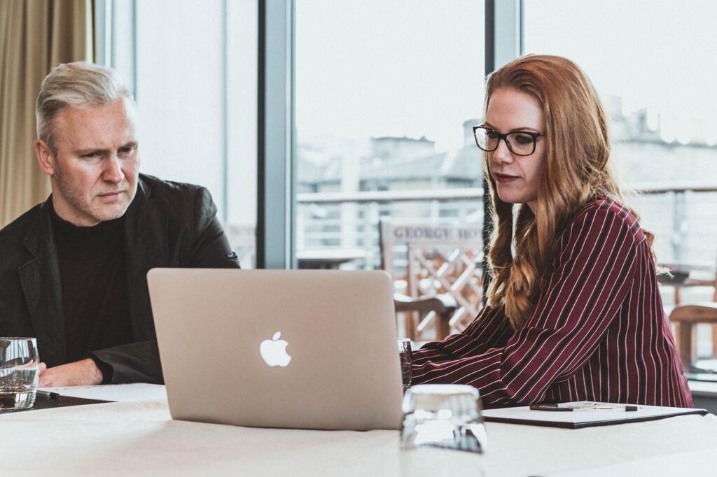 Two business professionals are sitting at a table, focused on a laptop, suggesting collaboration, online learning, or a work meeting in a modern office environment.
