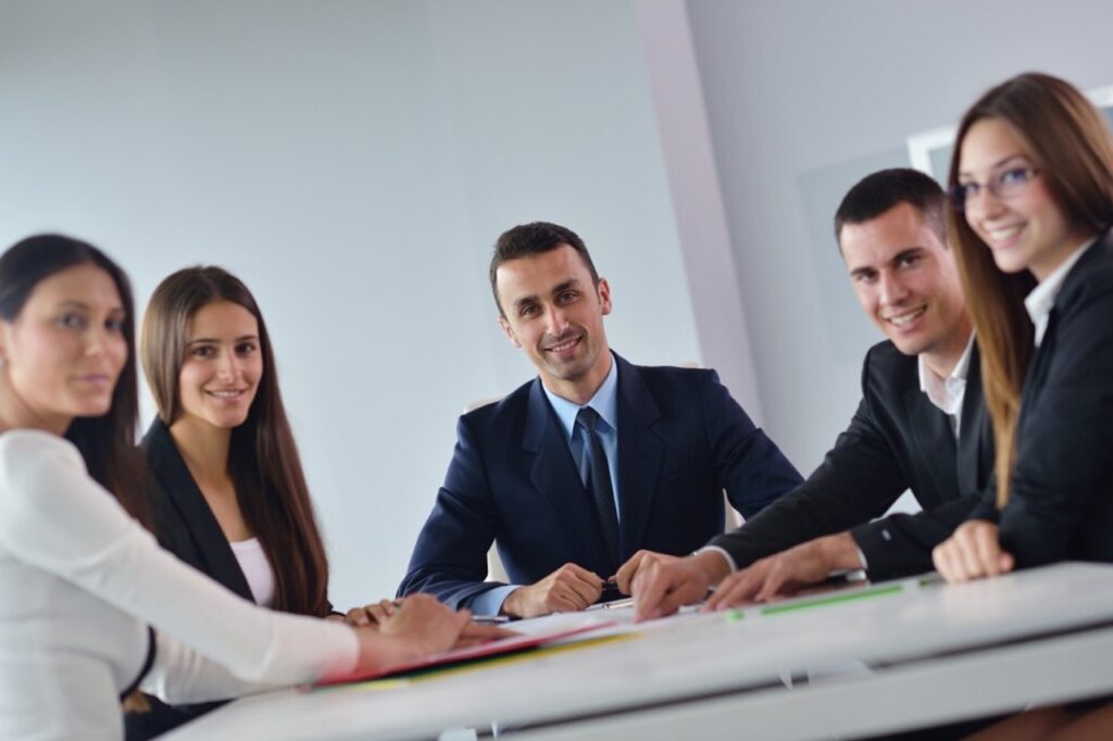 A group of business professionals in formal attire sit around a meeting table, smiling and engaged in discussion, suggesting teamwork and collaboration in a corporate setting.