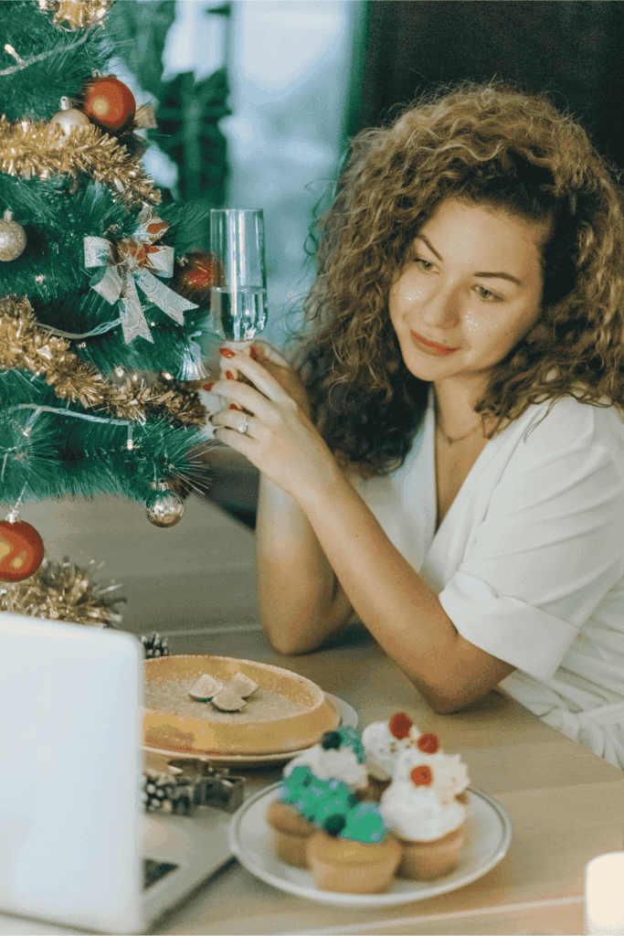 Woman celebrating Christmas with a drink beside a decorated tree and festive desserts.
