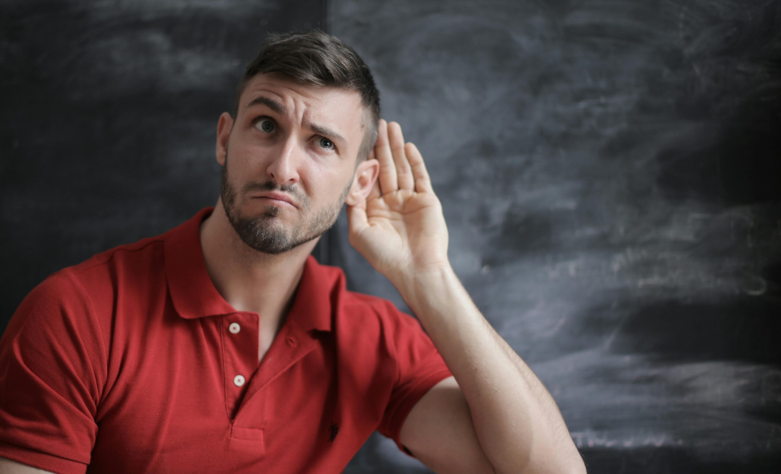 Man in a red shirt holding his hand to his ear as if listening closely.
