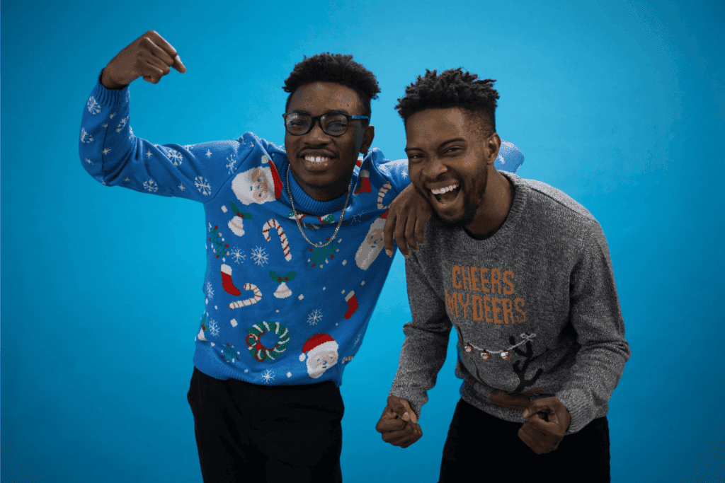 Two friends laughing and posing in festive Christmas sweaters against a blue background.
