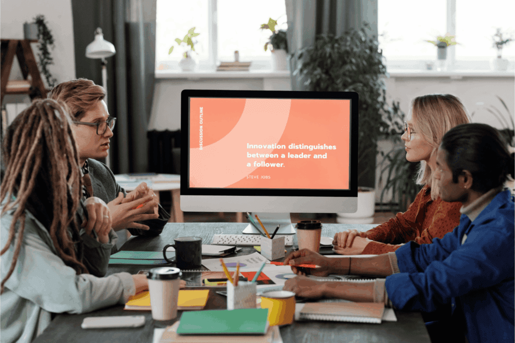 Group of colleagues in a meeting with a presentation screen displaying a Steve Jobs quote.
