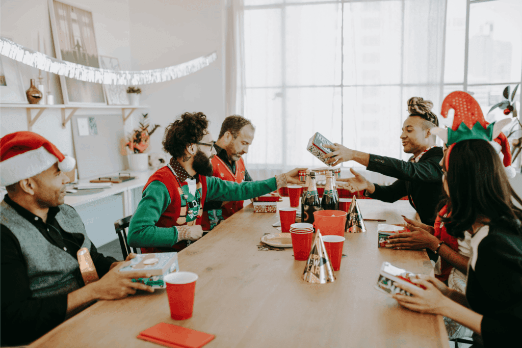 Coworkers celebrating a holiday party with gifts, festive hats, and decorations at the office.
