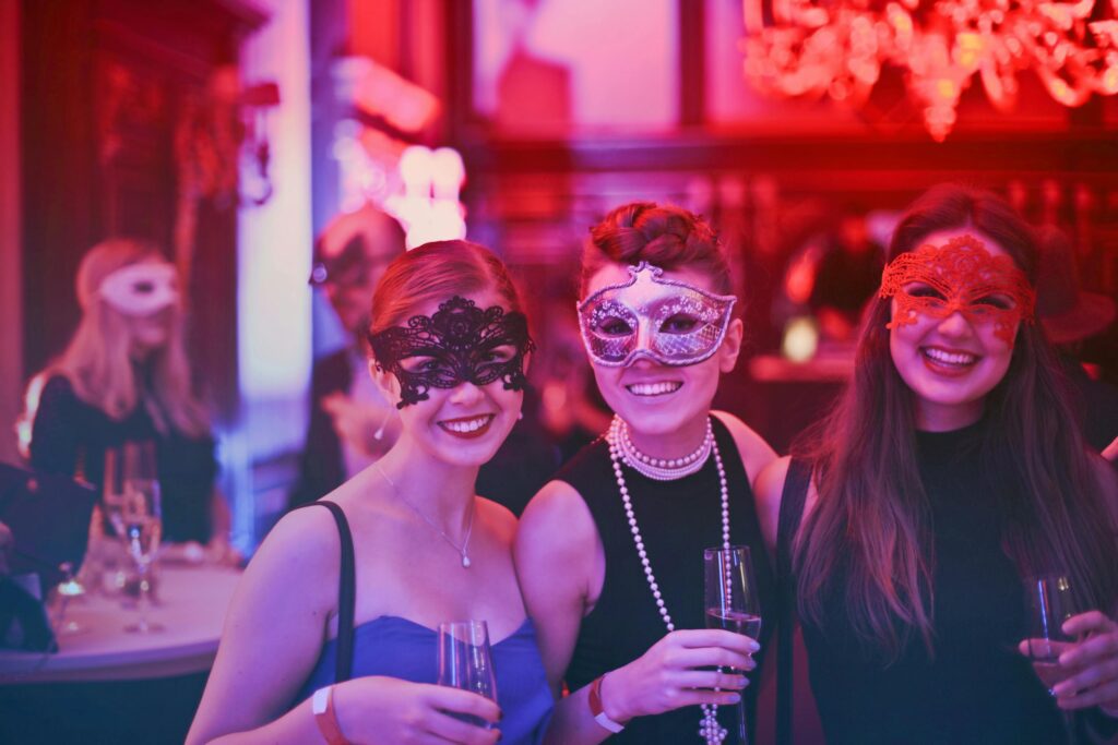Three women in elegant dresses wearing masquerade masks at a festive party.

